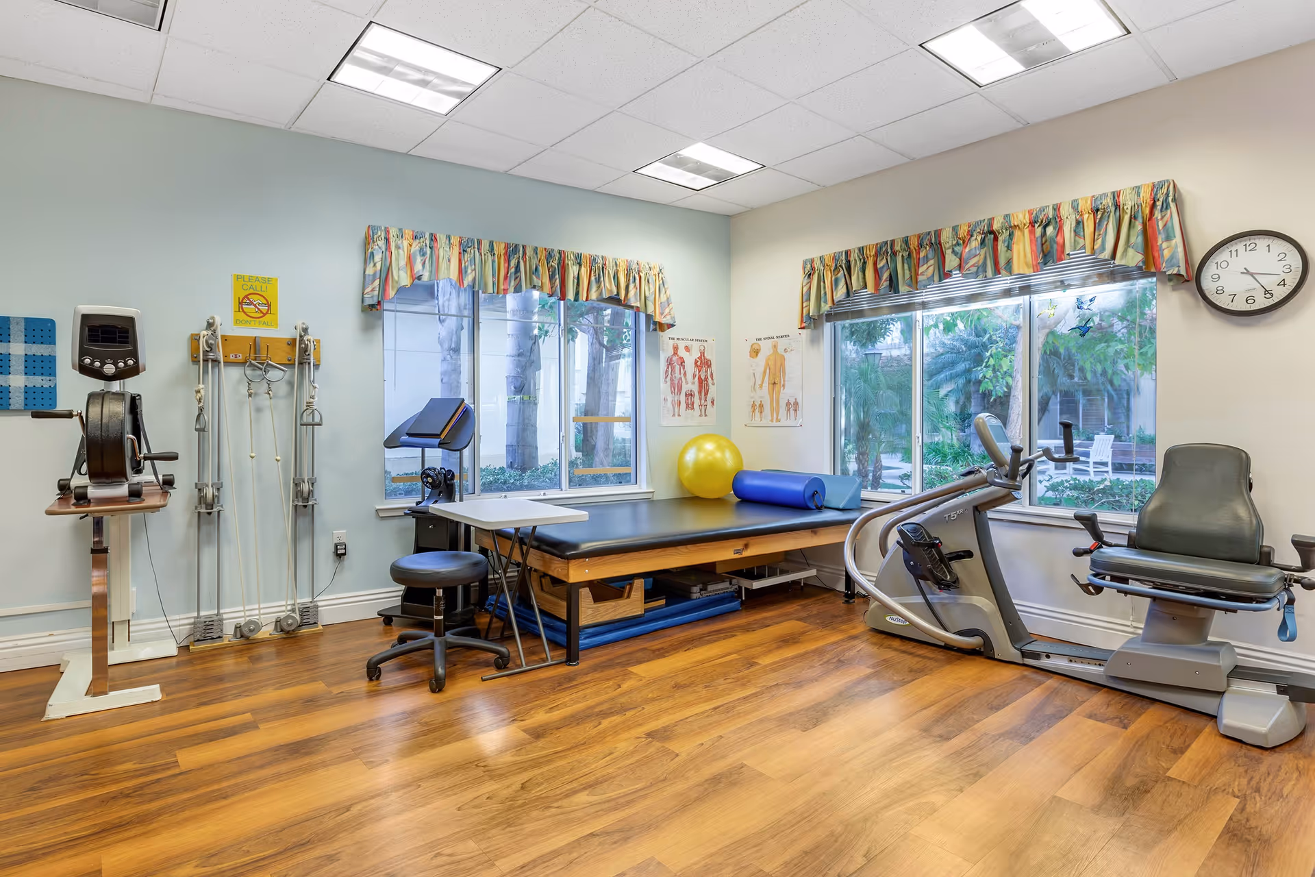 A senior living facility exercise room with wooden flooring, two large windows with colorful valances showing an outdoor garden. The room contains exercise equipment including a recumbent bike, a rowing machine, a therapy table with a yellow exercise ball and a blue foam roller, and a small stool. Anatomical charts are displayed on the walls and a clock shows the time as 3:40.