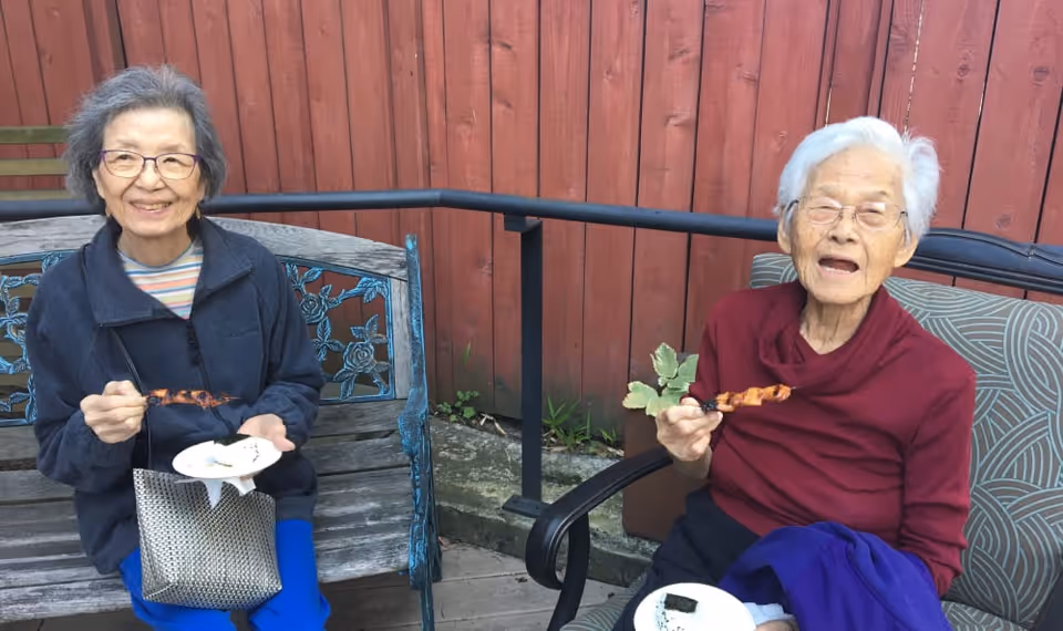 Two elderly women sitting outdoors on benches in front of a red wooden fence, smiling and holding plates with food and skewers.