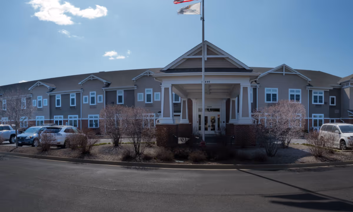 Front exterior view of a two-story senior living facility building with a covered entrance, several windows, parked cars, and a flagpole with flags against a blue sky with a few clouds.