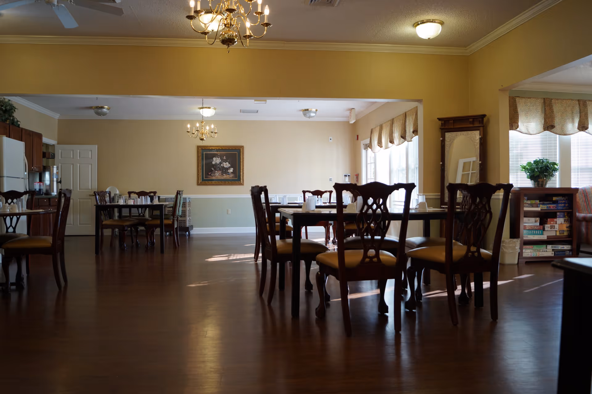 Spacious dining room with wooden tables and chairs, chandeliers, windows with valances, and a small bookshelf in the corner.