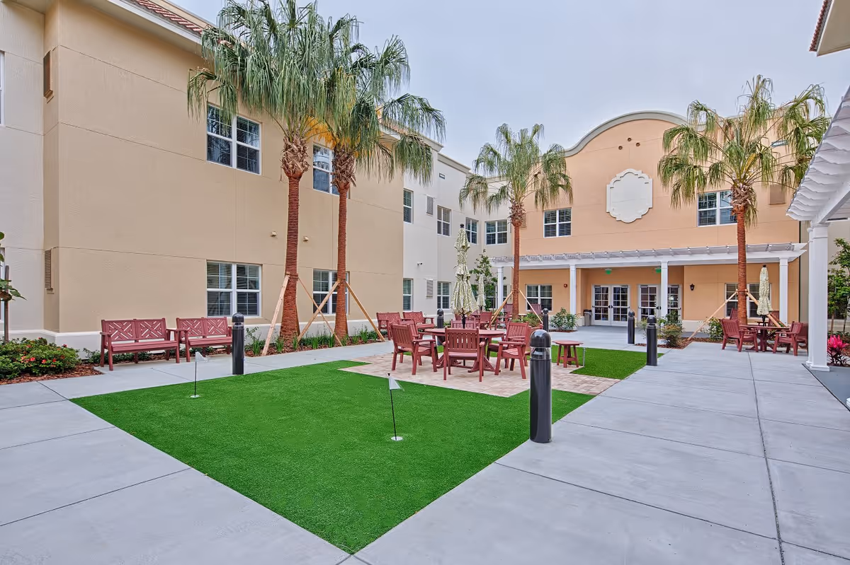 Outdoor courtyard area at Lexington Place featuring palm trees, red wooden benches and chairs arranged around tables with umbrellas, artificial putting green, and a beige building with multiple windows in the background.