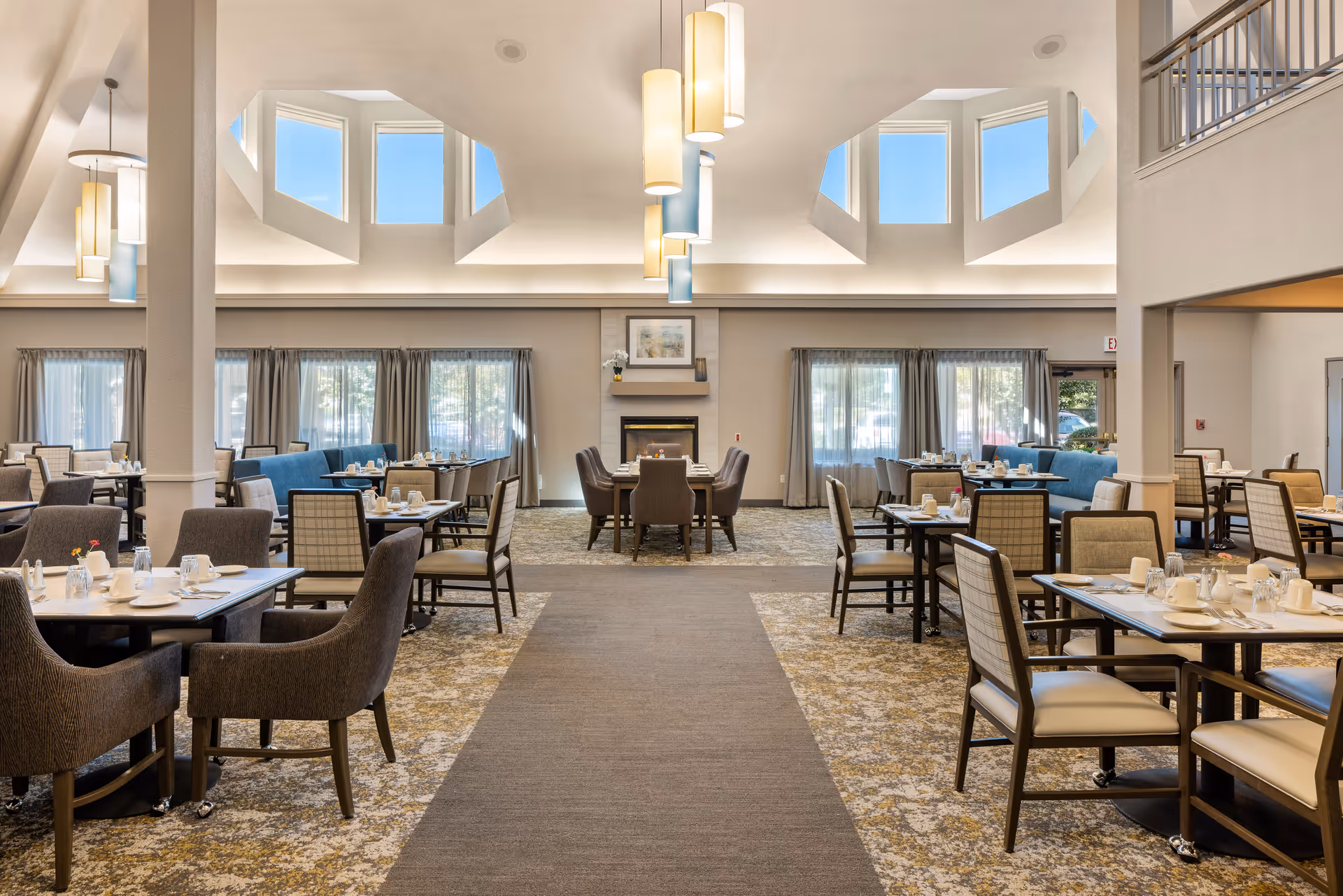 Spacious dining room in a senior living community with multiple tables and chairs arranged neatly. The room features large windows with curtains, high ceilings with modern pendant lights, and a central fireplace with artwork above it. The carpet has a patterned design, and the overall atmosphere is bright and welcoming.