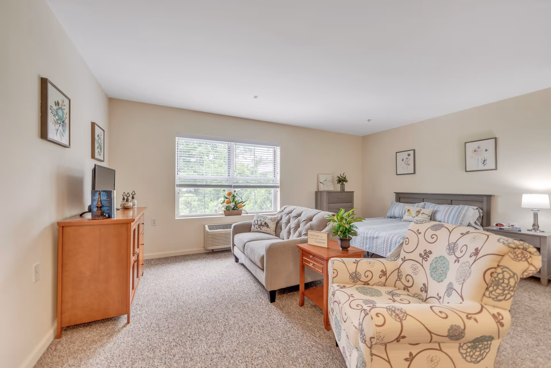 A bright and cozy senior living bedroom featuring a large window with blinds, a gray tufted sofa, a floral patterned armchair, a wooden TV stand with a flat-screen TV, a wooden side table with a potted plant, a bed with striped bedding, two nightstands with lamps, and framed artwork on the walls.
