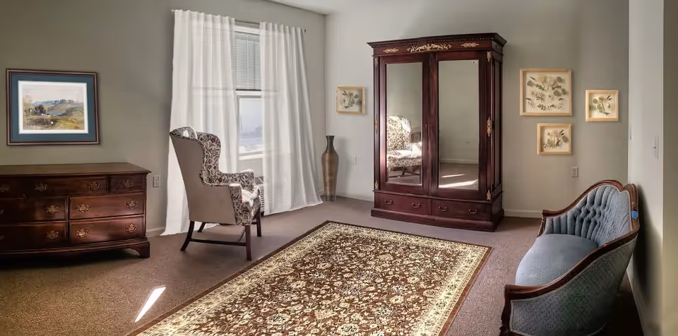 A cozy living room with classic wooden furniture including a large dresser, an ornate wardrobe with mirrored doors, a floral upholstered armchair, and a blue tufted loveseat. The room has beige walls, a window with white curtains, a decorative vase, framed botanical prints on the wall, and a patterned area rug on the carpeted floor.