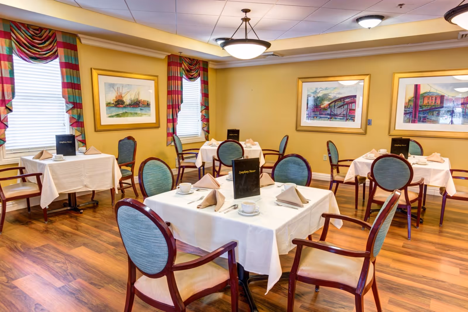 Bright dining room with multiple tables set with white tablecloths and place settings, upholstered chairs, framed artwork, and colorful curtains.