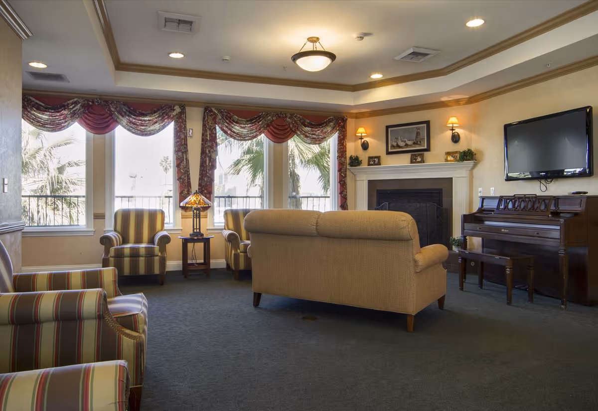 A cozy living room with a beige sofa and two striped armchairs facing a fireplace. There is a wooden piano with a bench on the right side and a flat-screen TV mounted on the wall above it. The room has large windows with floral and red valance curtains, allowing natural light to enter. A small table with a decorative lamp is positioned between the two armchairs near the windows. The ceiling has recessed lighting and a central light fixture.