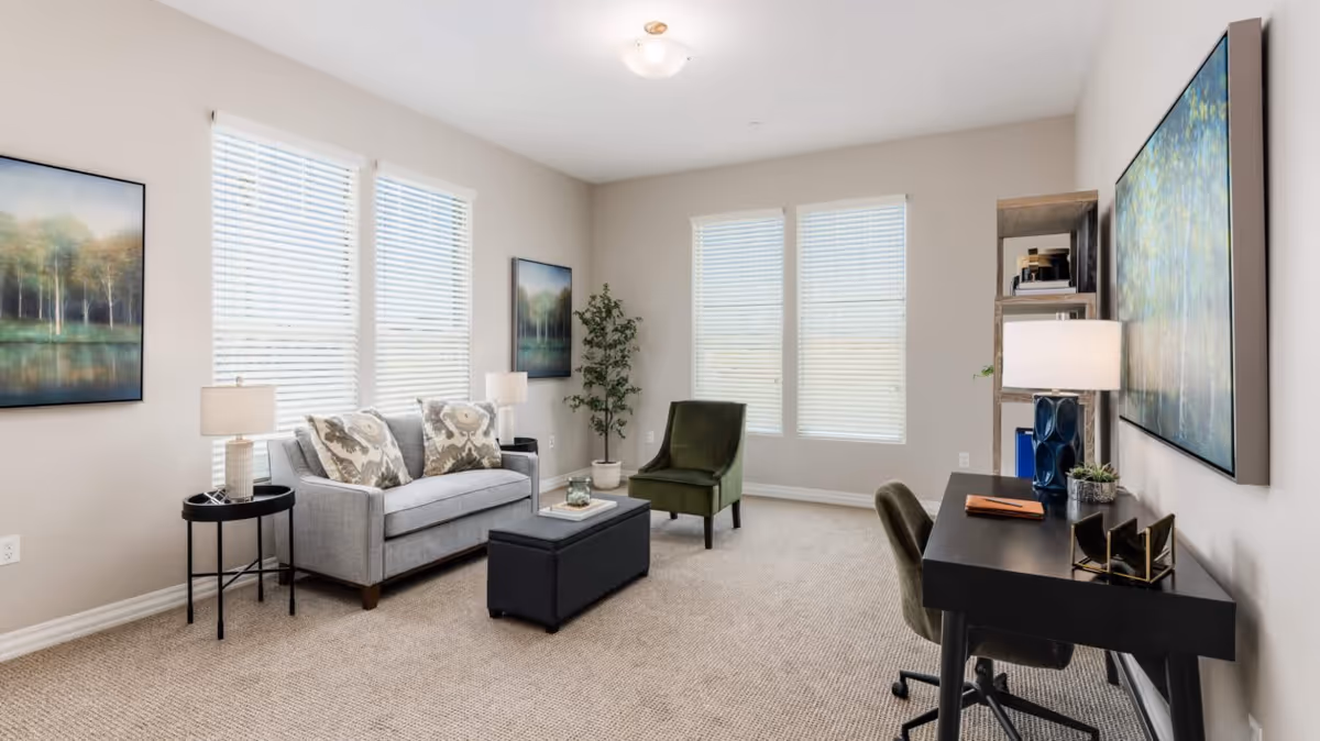 A bright and spacious living room with beige walls and carpeted floor. The room features a gray sofa with patterned cushions, a green armchair, a black ottoman coffee table, and a black desk with a green chair. There are two table lamps, a potted plant, and three large windows with white blinds. The walls are decorated with framed landscape paintings.