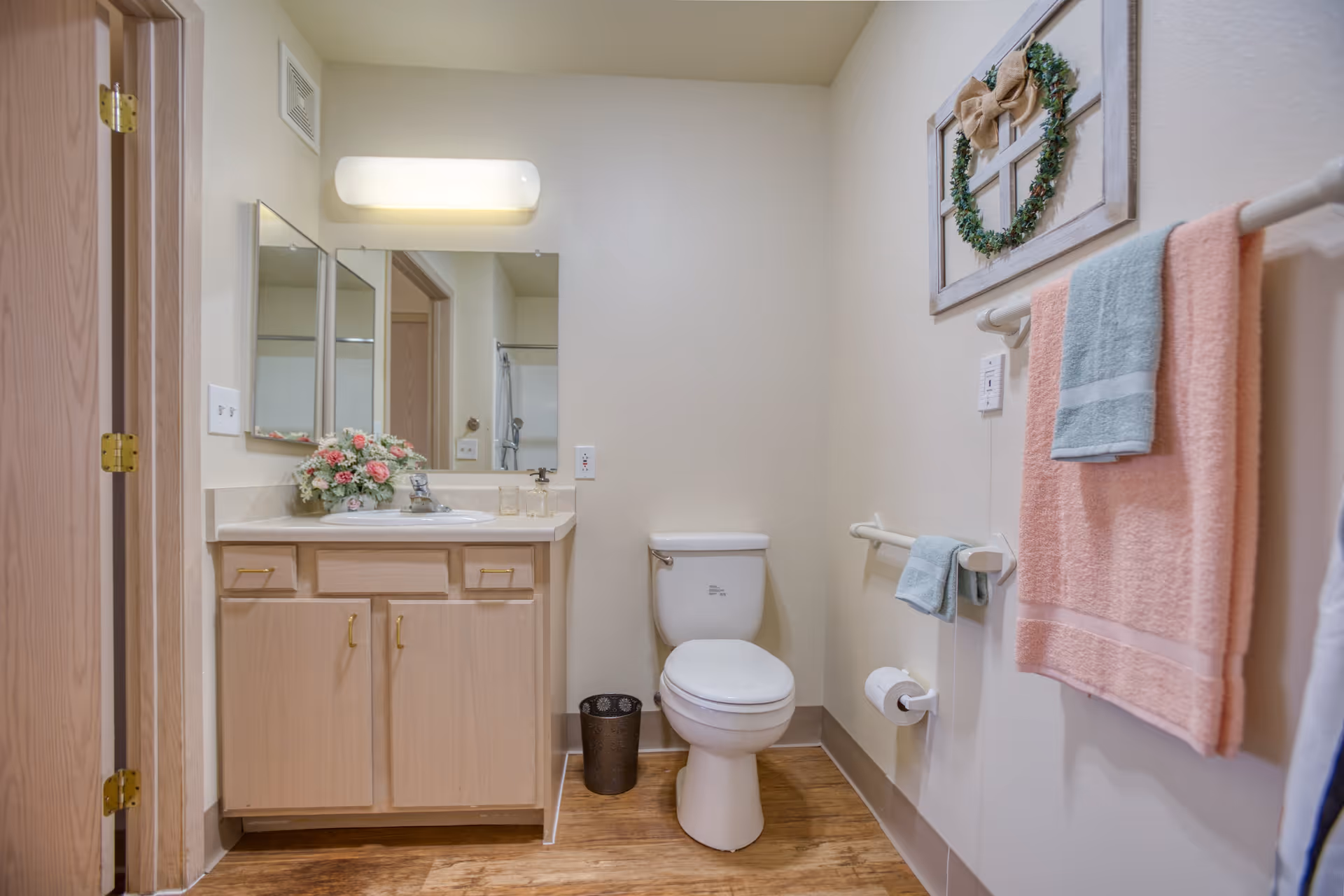 A clean and well-lit bathroom featuring a white toilet, a wooden vanity with a sink, a mirror above the sink, and a floral arrangement on the countertop. There are pastel-colored towels hanging on a towel rack on the right wall, and a decorative wreath hanging above the towels. The floor has a wood-like finish.