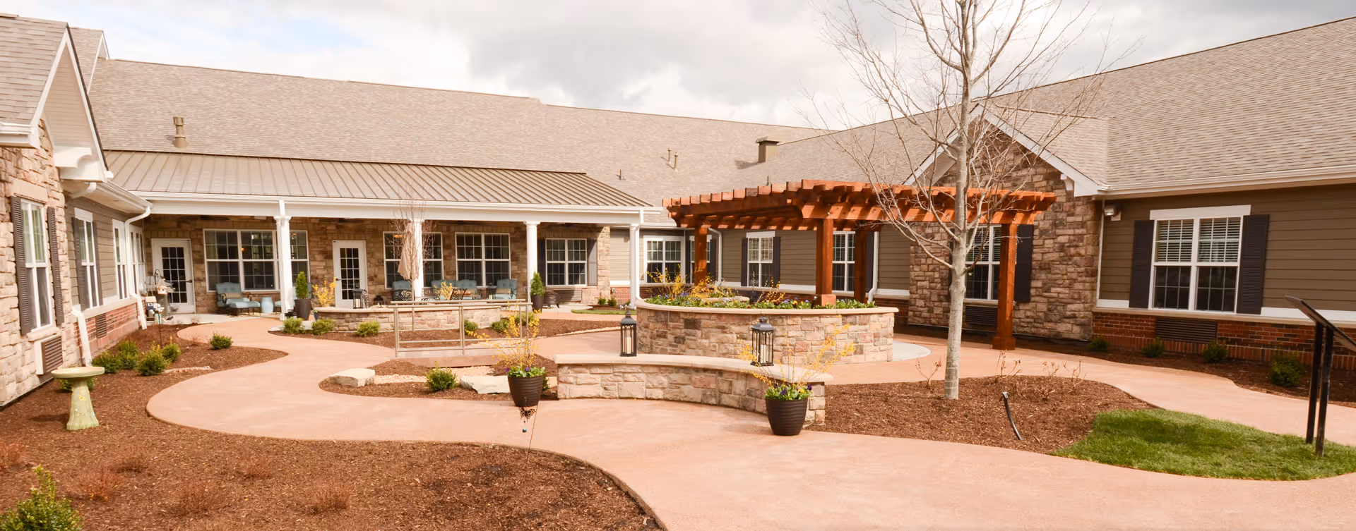 Outdoor courtyard area of a senior living facility with a paved walkway, a wooden pergola over a circular stone planter, several potted plants, and seating areas along the building's exterior walls. The building has stone and siding walls with multiple windows and doors.