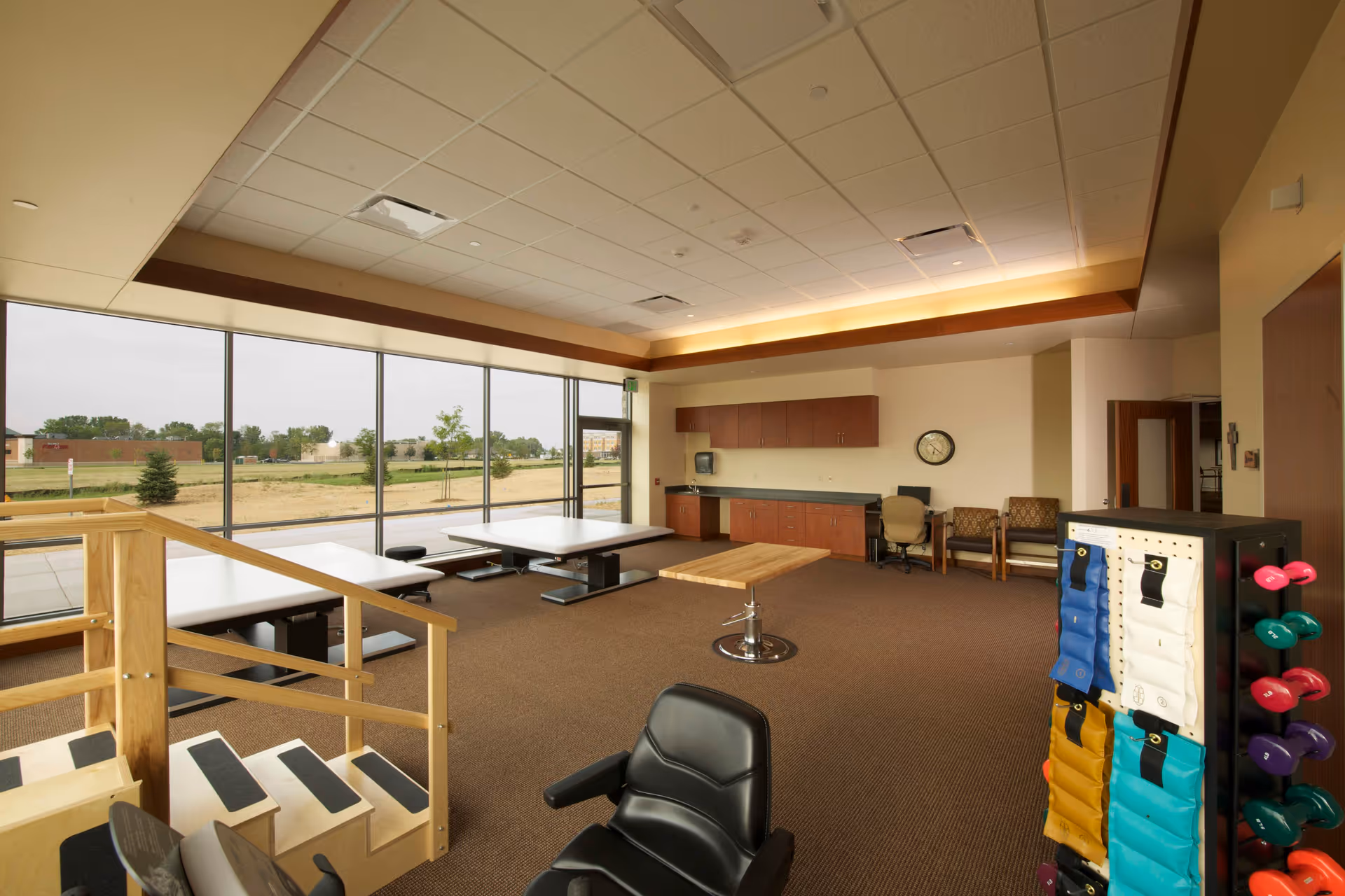 Bright rehabilitation/exercise room with floor-to-ceiling windows, therapy tables, a small set of stairs, cabinets, seating and a rack of colorful weights.