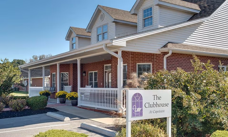 Exterior view of a red brick building with white trim and a covered porch with white railings. There are potted plants with yellow flowers on the porch and a sign in front that reads 'The Clubhouse At Capetown'. The sky is clear and blue.