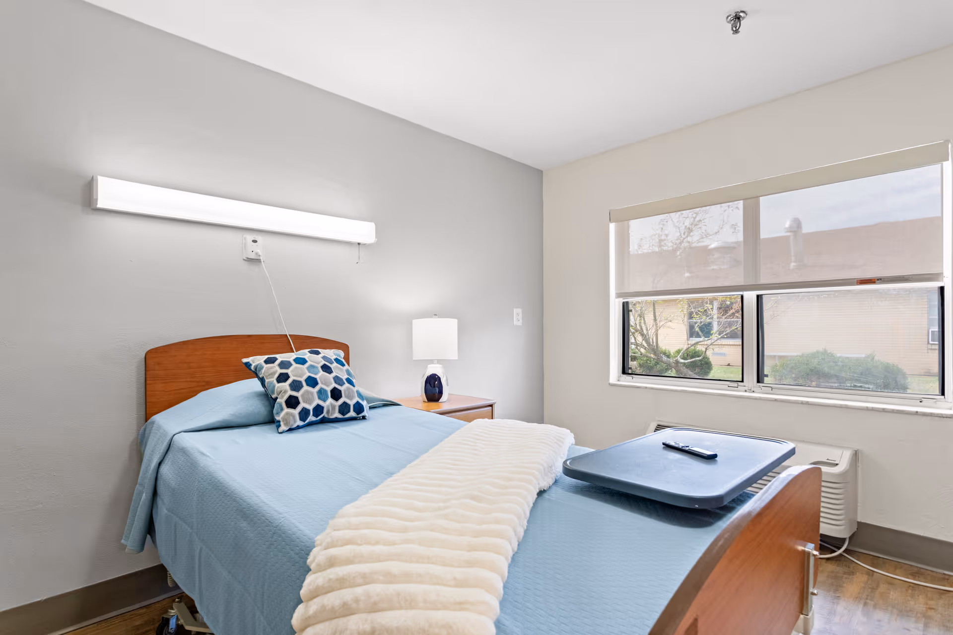 A simple, clean nursing care bedroom with a single bed covered in a light blue blanket and a white throw. There is a hexagon-patterned pillow on the bed, a bedside table with a lamp, a large window with a roller shade partially down, and a tray with a remote control on the bed. The walls are painted light gray and white.