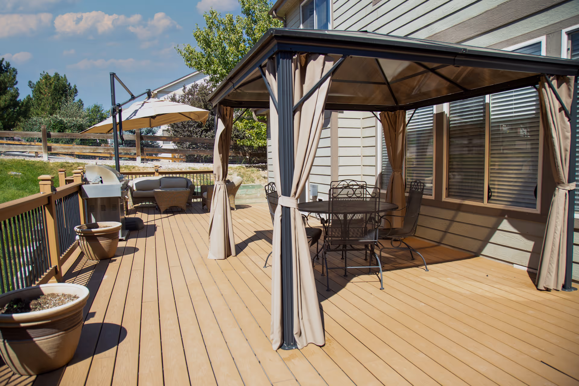 Outdoor wooden deck area with a metal gazebo featuring a round table and four chairs. There are potted plants along the railing, a barbecue grill, and a seating area with a sofa and umbrella. The deck is attached to a light-colored building with windows and surrounded by greenery and a wooden fence.