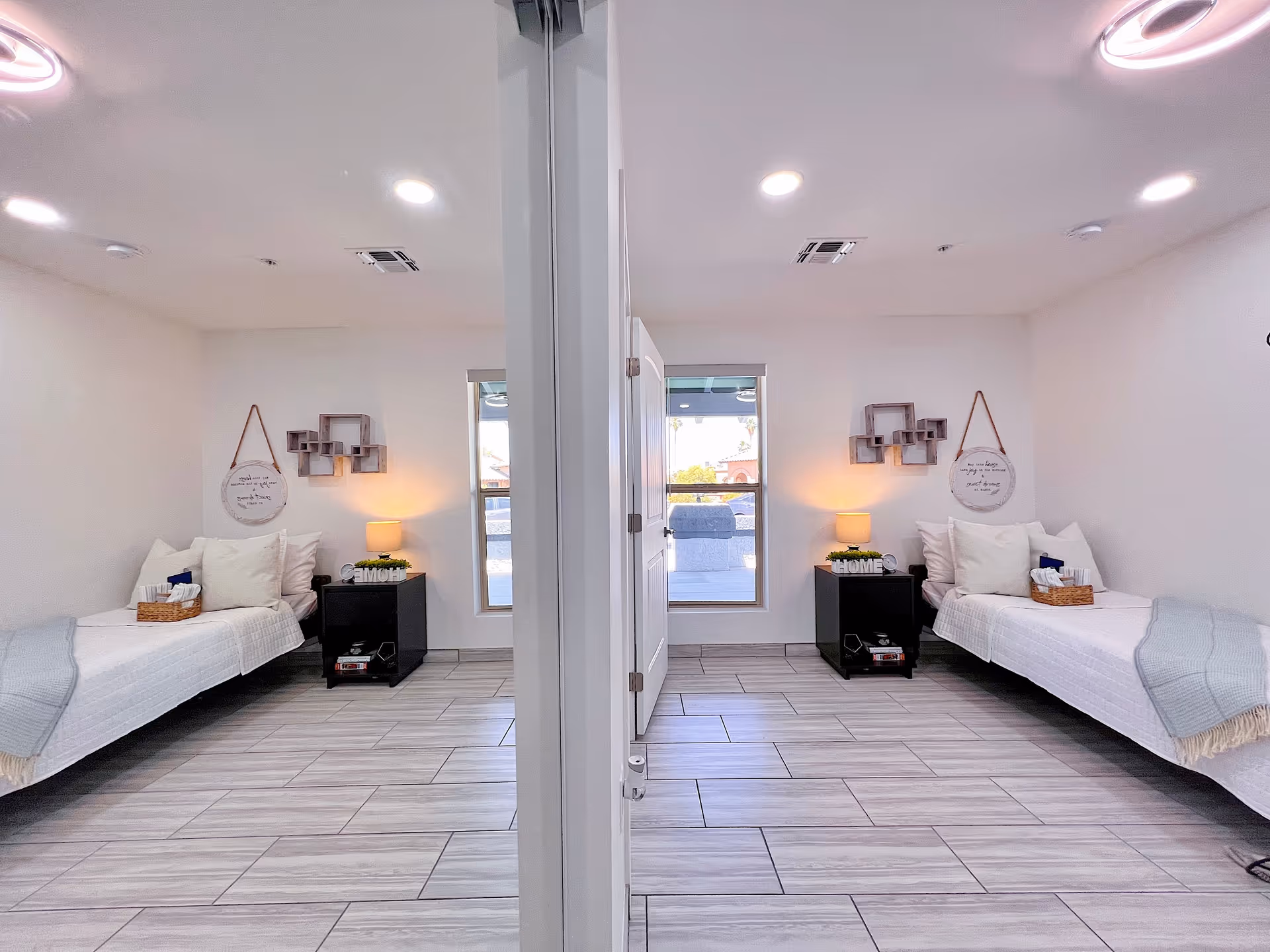 A bright, clean bedroom with two single beds separated by a partial wall. Each bed is neatly made with white bedding, pillows, and a light blue throw blanket. Beside each bed is a black nightstand with a small lamp, decorative items, and a basket with tissues. The room has light-colored tiled flooring and white walls with minimal decor, including a round hanging sign and a geometric wall shelf. A window at the far end lets in natural light.