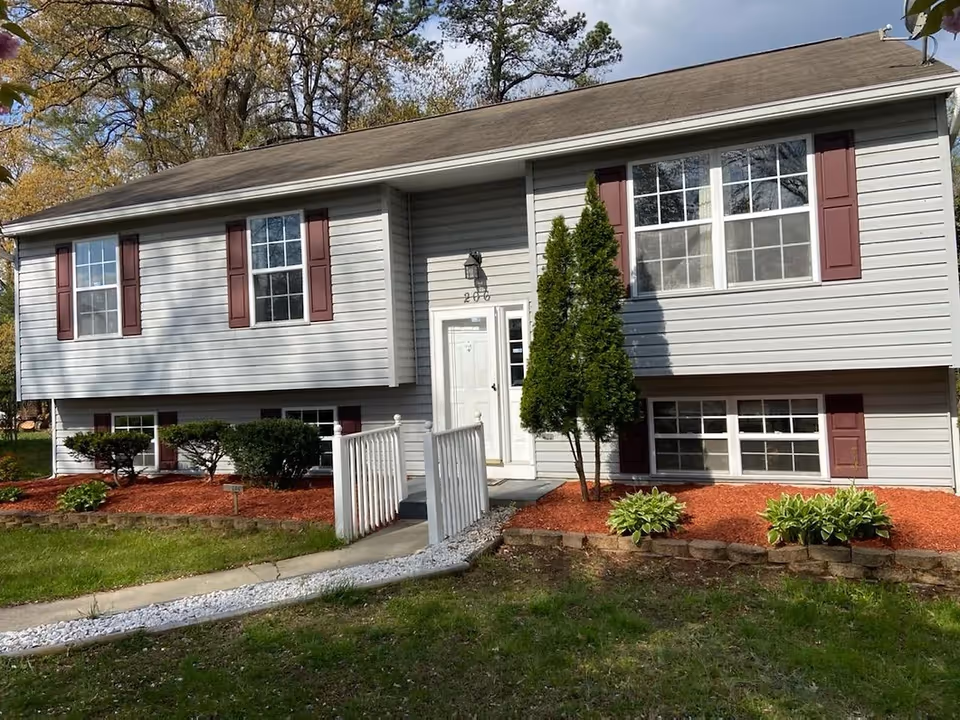 Front exterior of a light-gray split-level house with maroon shutters, a white entry ramp, and landscaped red-mulch beds.