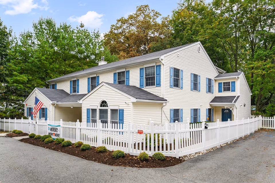 White, two-story memory care building with blue shutters and a white picket fence.