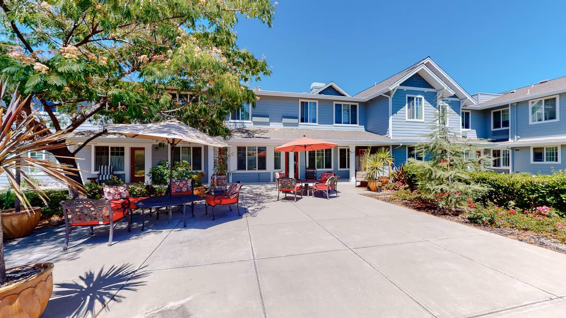 Outdoor patio area at The Commons At Dallas Ranch featuring seating arrangements with red and floral cushions under large umbrellas, surrounded by potted plants and trees, with a two-story blue and white building in the background under a clear blue sky.
