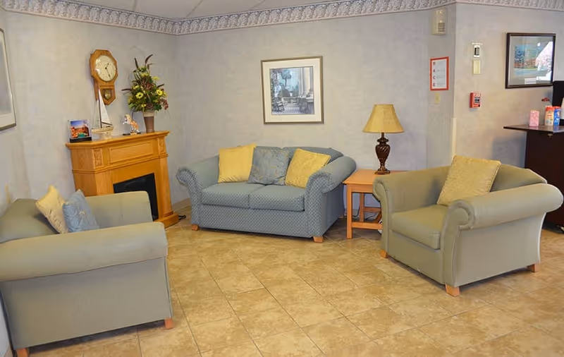 A cozy living room area with three upholstered armchairs and a loveseat, all with cushions. There is a wooden side table with a lamp, a wooden mantel with a clock and flower arrangement, and framed artwork on the walls. The floor is tiled and the walls have a light-colored wallpaper with a decorative border near the ceiling.