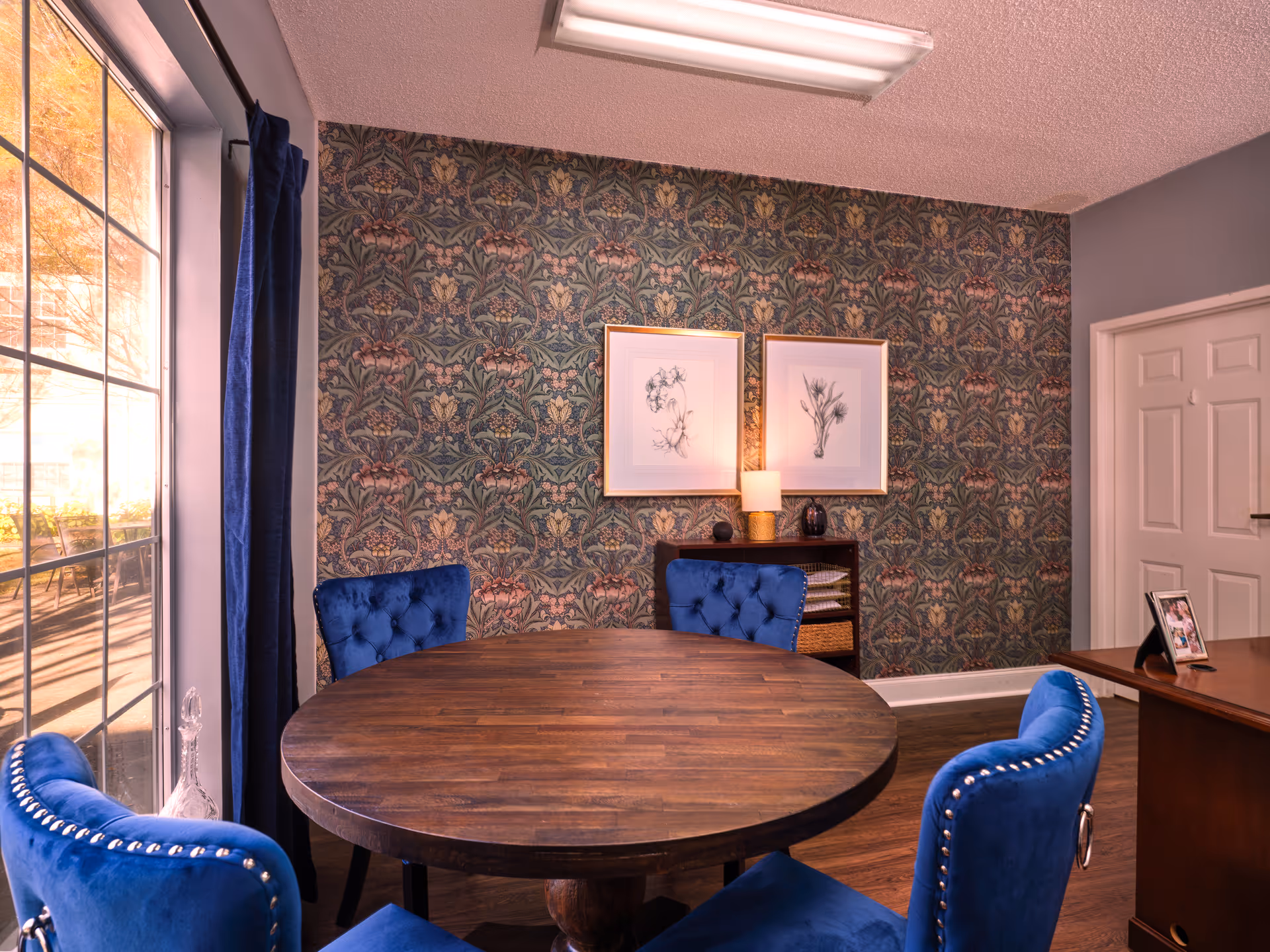 A cozy interior room featuring a round wooden table surrounded by four blue upholstered chairs with silver nailhead trim. The room has a large window with blue curtains allowing natural light to enter. The wall behind the table is decorated with patterned wallpaper and two framed botanical prints. A small wooden shelf with a lamp and decorative items is placed against the wall. A white door and part of a wooden desk with a photo frame are visible on the right side.