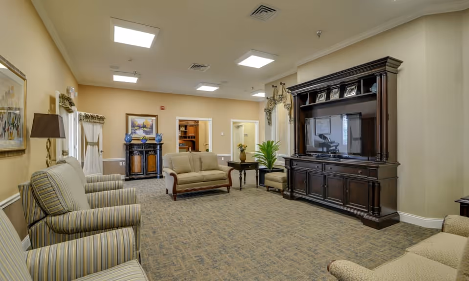 A spacious senior living facility common area with beige walls and carpeted floor. The room features several upholstered armchairs and sofas arranged around a large dark wood entertainment center with a flat-screen TV. Decorative elements include framed paintings, a table with a vase of flowers, and a potted plant. The ceiling has recessed lighting and air vents.