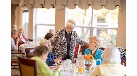 A group of elderly residents socializing and dining together in a bright dining room, with one woman standing and greeting others seated at a table.