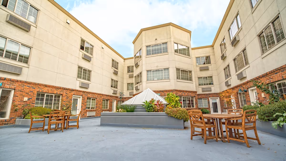 Outdoor courtyard area of a senior living facility with wooden tables and chairs, surrounded by a three-story building with multiple windows and some greenery in planters.