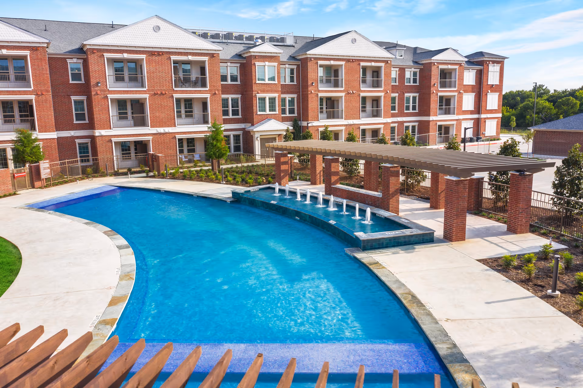 Exterior view of a multi-story brick senior living building with balconies, a curved outdoor swimming pool, and a pergola-covered patio.