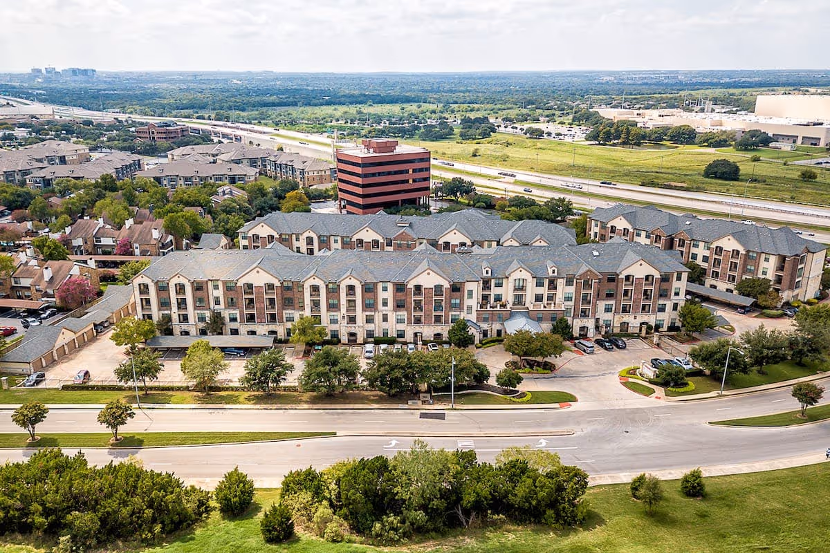 Aerial view of a multi-story residential building complex surrounded by trees, roads, and parking areas, with a highway and green fields in the background under a partly cloudy sky.
