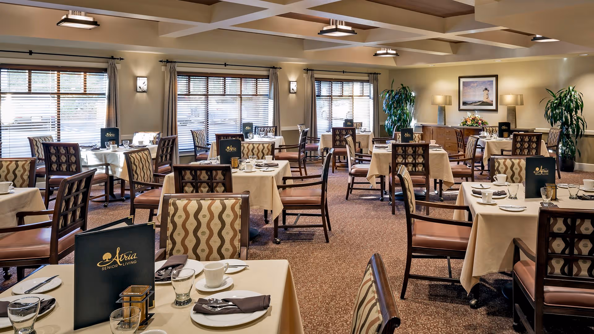 A well-lit dining room in a senior living facility with multiple tables covered in beige tablecloths, each set with cups, glasses, napkins, and menus labeled 'Atria Senior Living'. The room features patterned chairs, large windows with blinds, potted plants, and framed artwork on the walls.