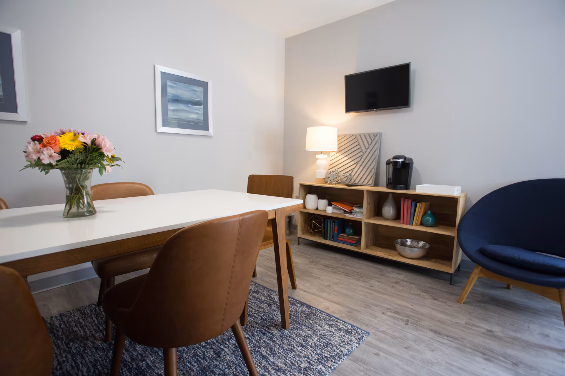A modern dining room with a white rectangular table surrounded by brown leather chairs. A vase with colorful flowers is placed on the table. On the right side, there is a wooden shelving unit with books, decorative items, a lamp, and a coffee maker. Above the shelving unit, a flat-screen TV is mounted on the wall. A blue cushioned chair is positioned to the right of the shelving unit. The room has light gray walls, wooden flooring, and a blue patterned rug under the table.