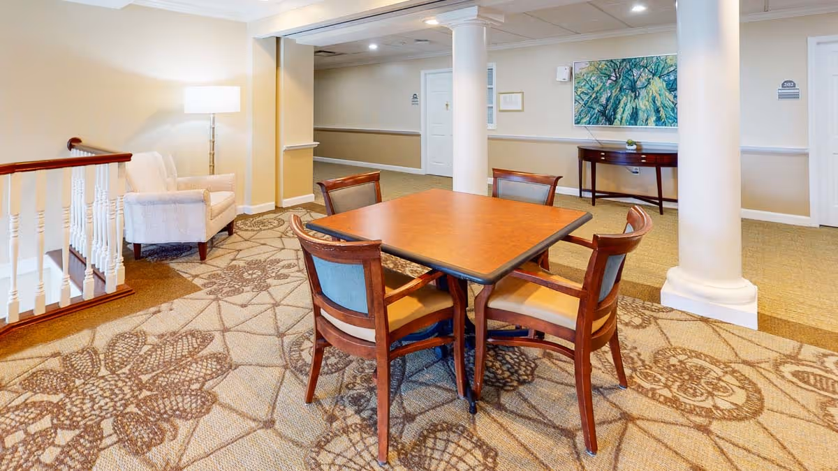 Carpeted communal lounge area featuring a square wooden table with four chairs, columns, an armchair, and artwork on the wall.