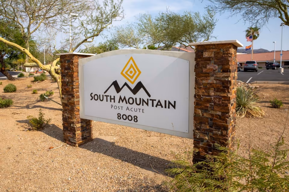 Stone-framed outdoor sign reading "South Mountain Post Acute 8008" in a landscaped desert front with a parking lot and flags behind it.