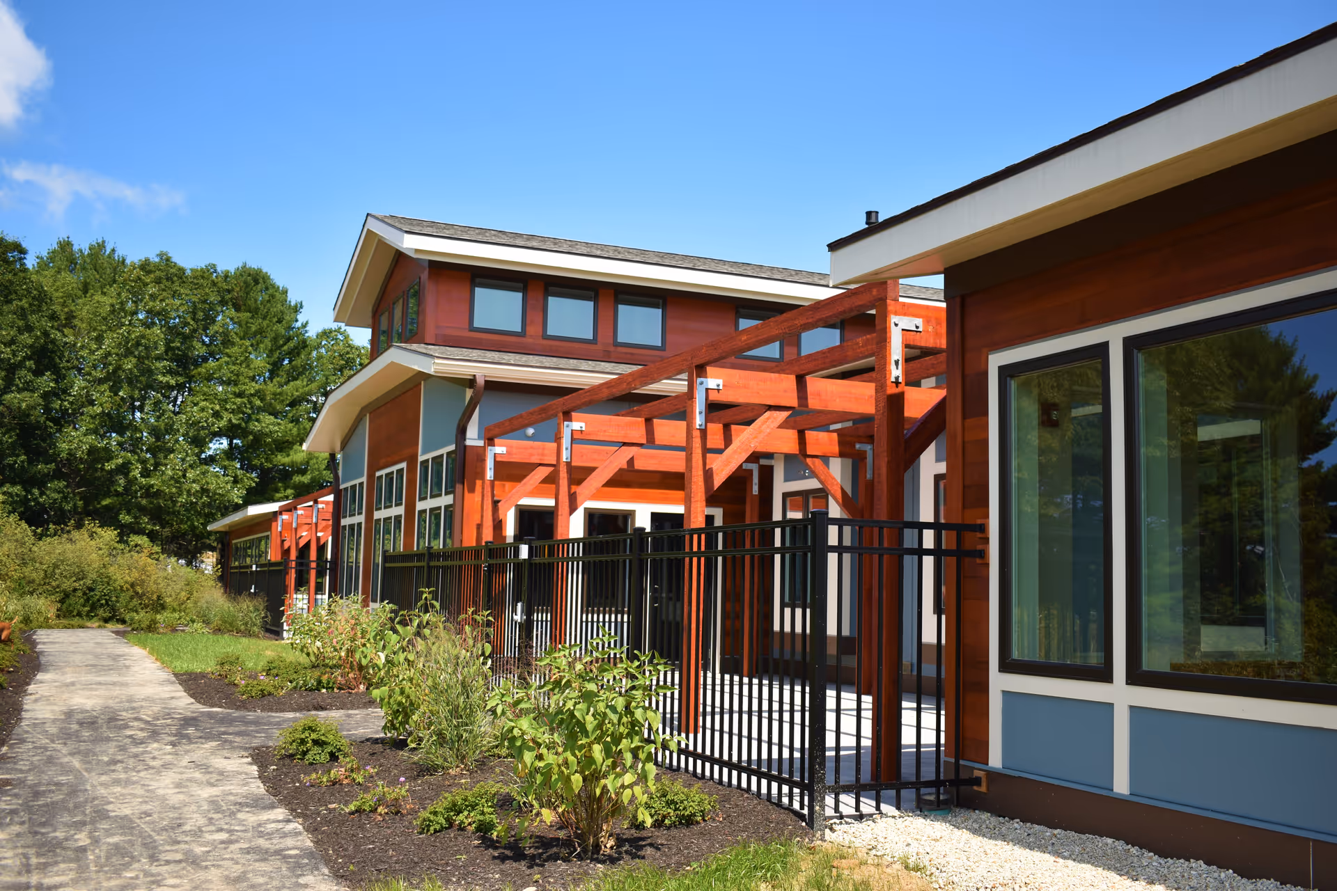 Exterior view of a modern senior living facility building with wooden accents and large windows, a black metal fence, a paved walkway, and landscaped greenery under a clear blue sky.
