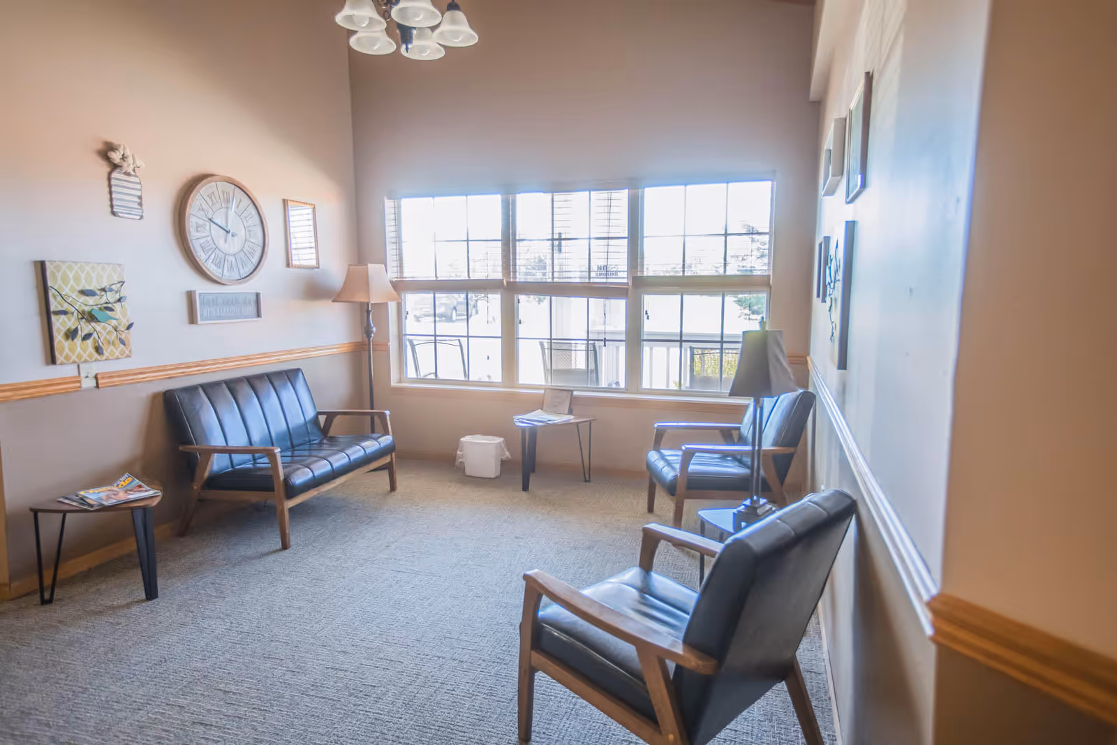 A cozy sitting area with three black leather chairs and a matching loveseat arranged around a small table. The room has beige walls adorned with decorative wall art and a large clock. A floor lamp and a table lamp provide lighting, and a large window lets in natural light, showing an outdoor view.