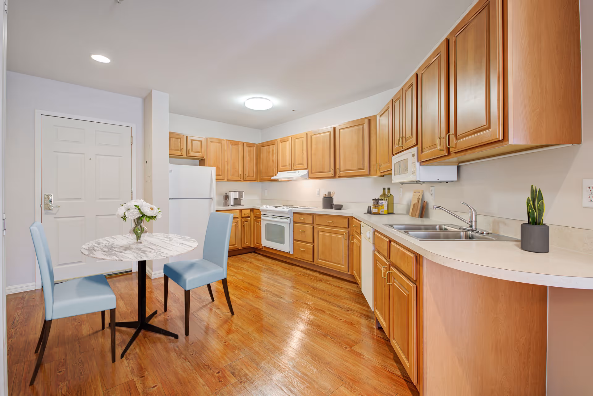 A bright kitchen with wooden cabinets, a white refrigerator, stove, microwave, and dishwasher. There is a small round table with a marble-like top and two light blue chairs. A vase with white flowers is on the table, and a small potted plant is on the countertop near the sink. The floor is wooden, and the walls are light-colored.