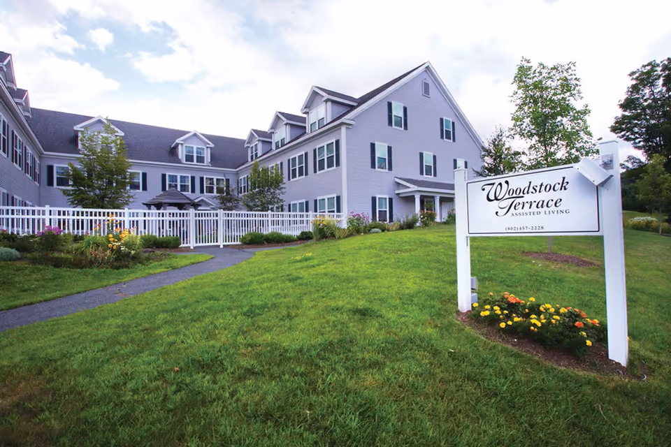 Exterior view of Woodstock Terrace assisted living facility showing a large, light gray building with multiple windows and dormers. The building is surrounded by a white fence, green lawn, trees, and flower beds. A white sign with the facility name and contact number is visible in the foreground.