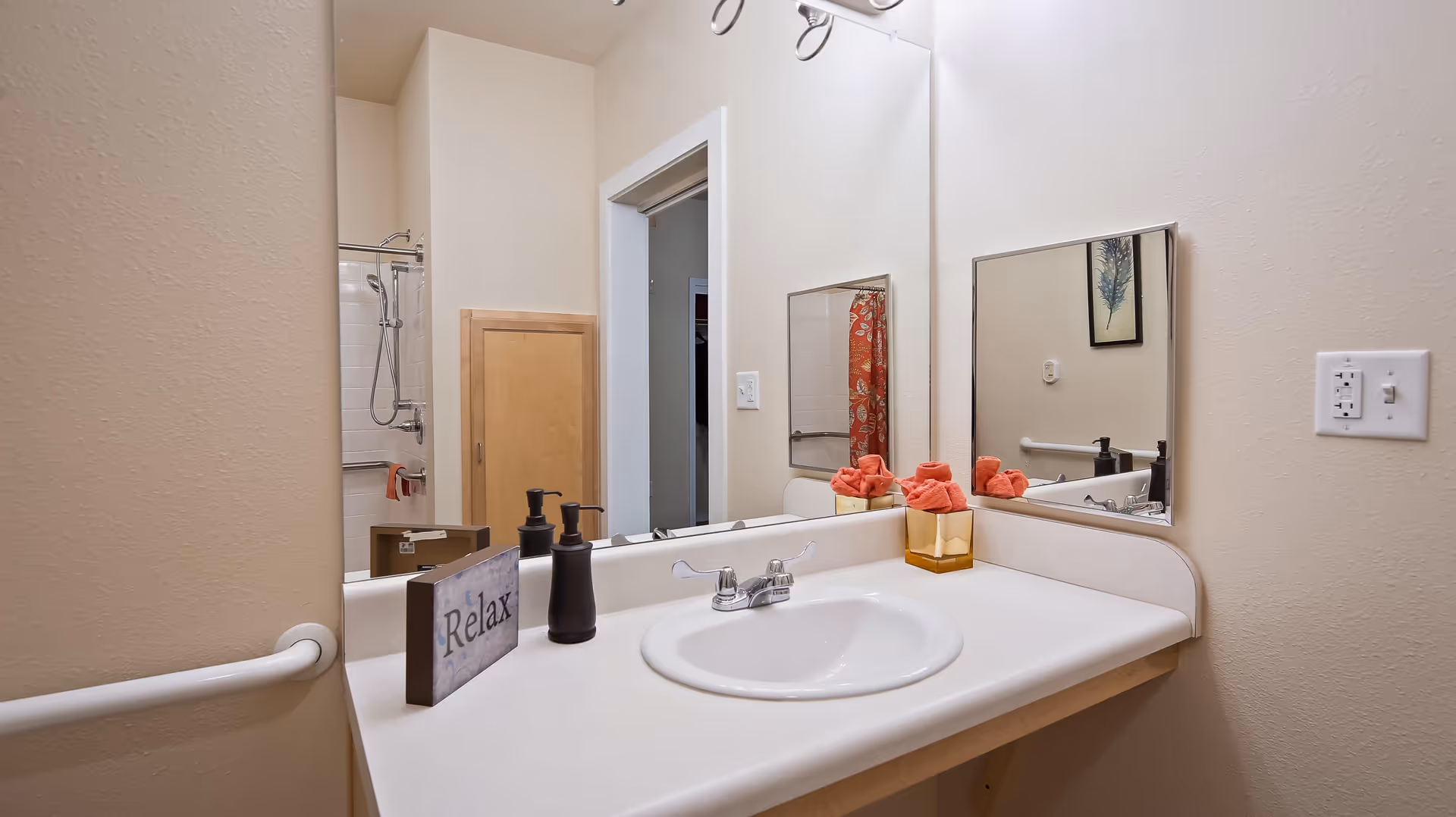 A bathroom sink area with a white countertop, a silver faucet, and a large mirror above. On the countertop, there is a small decorative sign that says 'Relax', two black soap dispensers, and a container holding rolled orange towels. In the reflection of the mirror, a shower with a handheld showerhead and a red shower curtain is visible. The walls are painted beige, and there is a white grab bar on the left side of the sink.