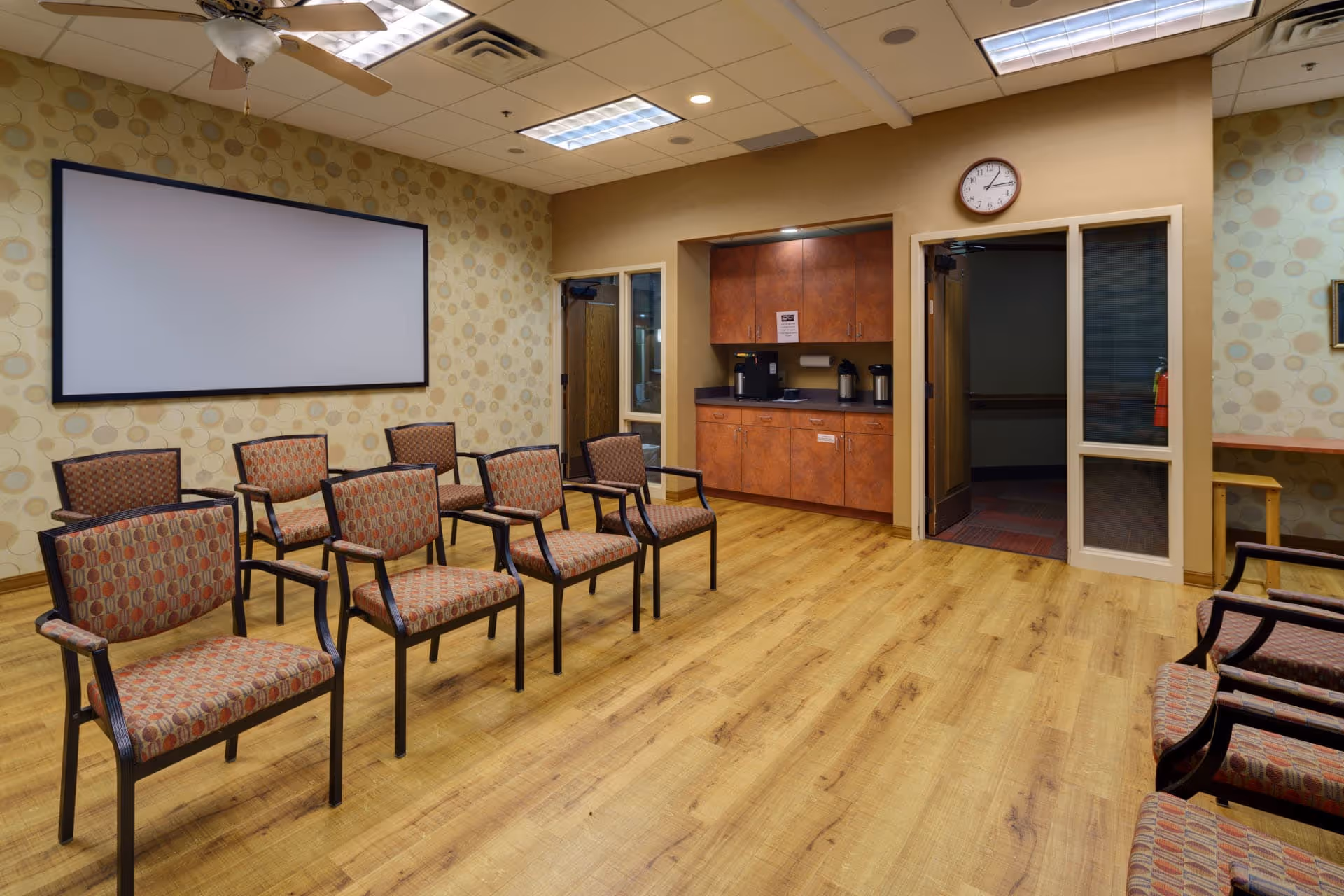 Meeting/activity room with rows of patterned chairs facing a large blank screen, wood floors, and a small kitchenette area.