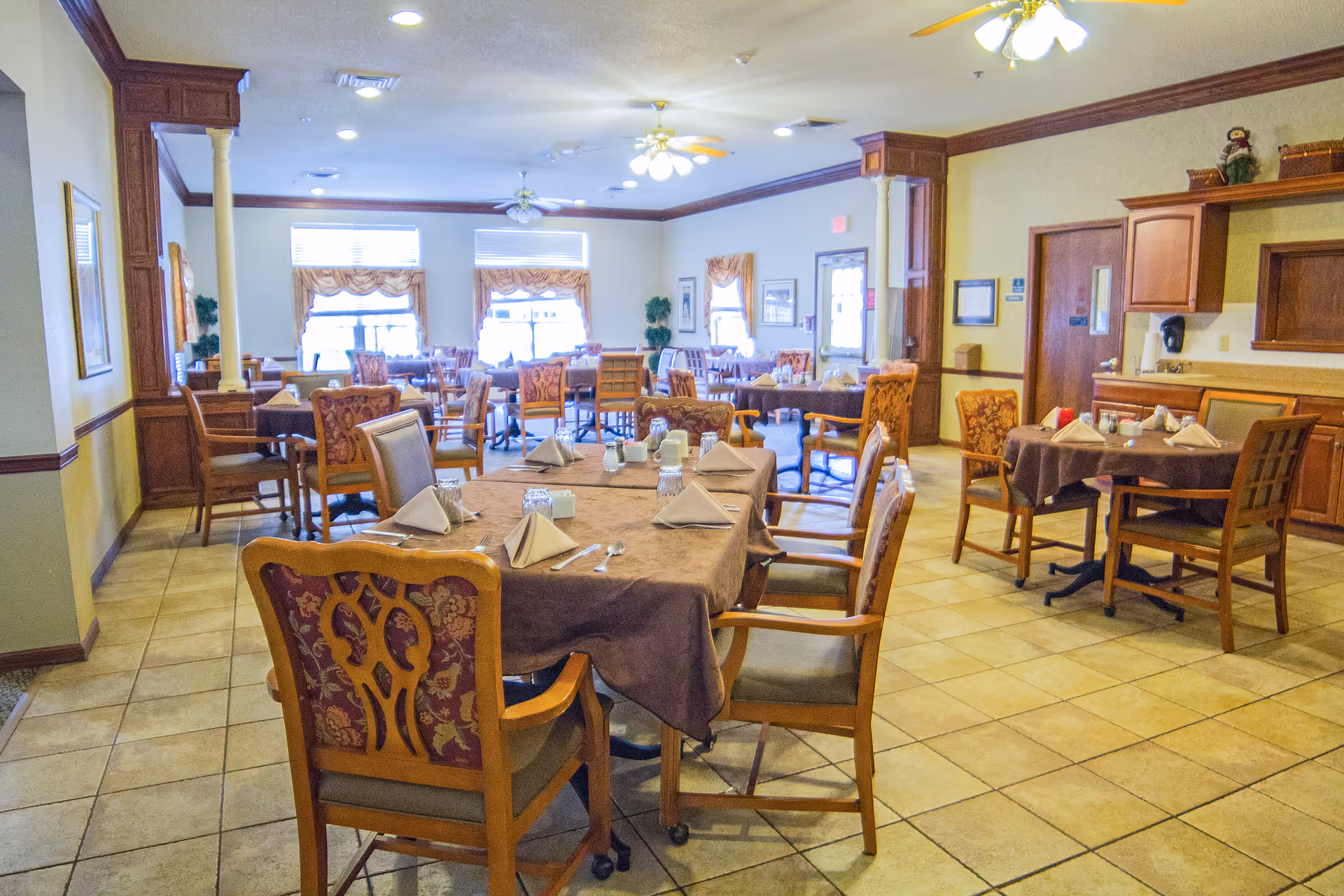 Dining room interior with multiple set tables and chairs, warm wood trim, and tiled floor in a senior living facility.