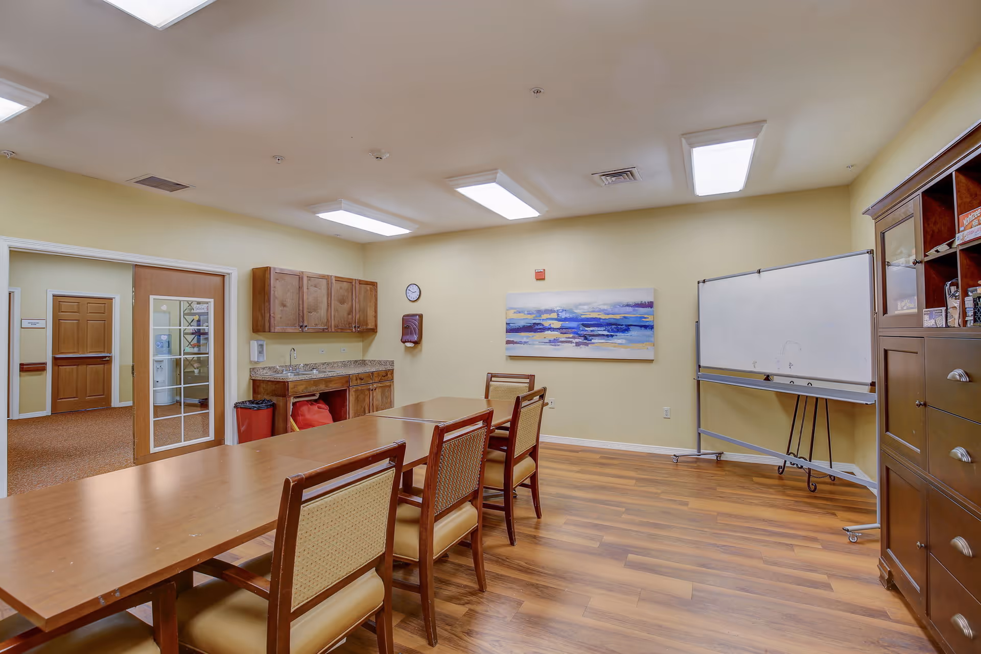 A meeting or activity room with a long wooden table surrounded by chairs, a whiteboard on wheels, a wooden cabinet with shelves containing board games, and a countertop with cabinets above and below. The room has wood flooring, beige walls, and fluorescent ceiling lights. A colorful abstract painting hangs on the wall.