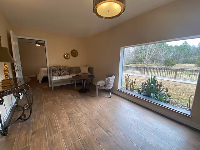 A cozy sitting area with a cushioned bench, a small round table, and an upholstered chair next to a large window overlooking an outdoor garden with plants and trees. The room has wood-look tile flooring, a decorative wall with two round wall hangings, and a ceiling light fixture. A doorway leads to a bedroom with a visible bed.