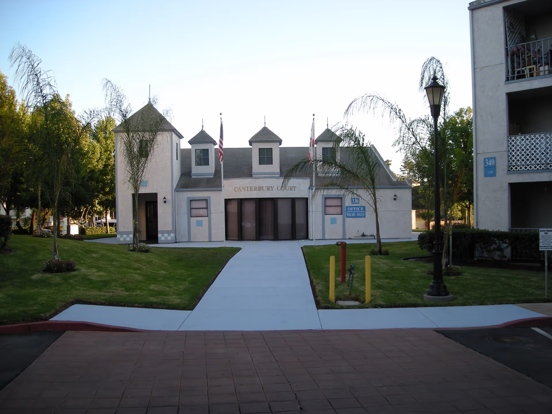 Exterior view of Canterbury Court Senior Apartments office building with a paved walkway leading to the entrance, flanked by small palm trees and green lawns. Two flagpoles with flags stand in front of the building, which has a light-colored facade and multiple small windows.