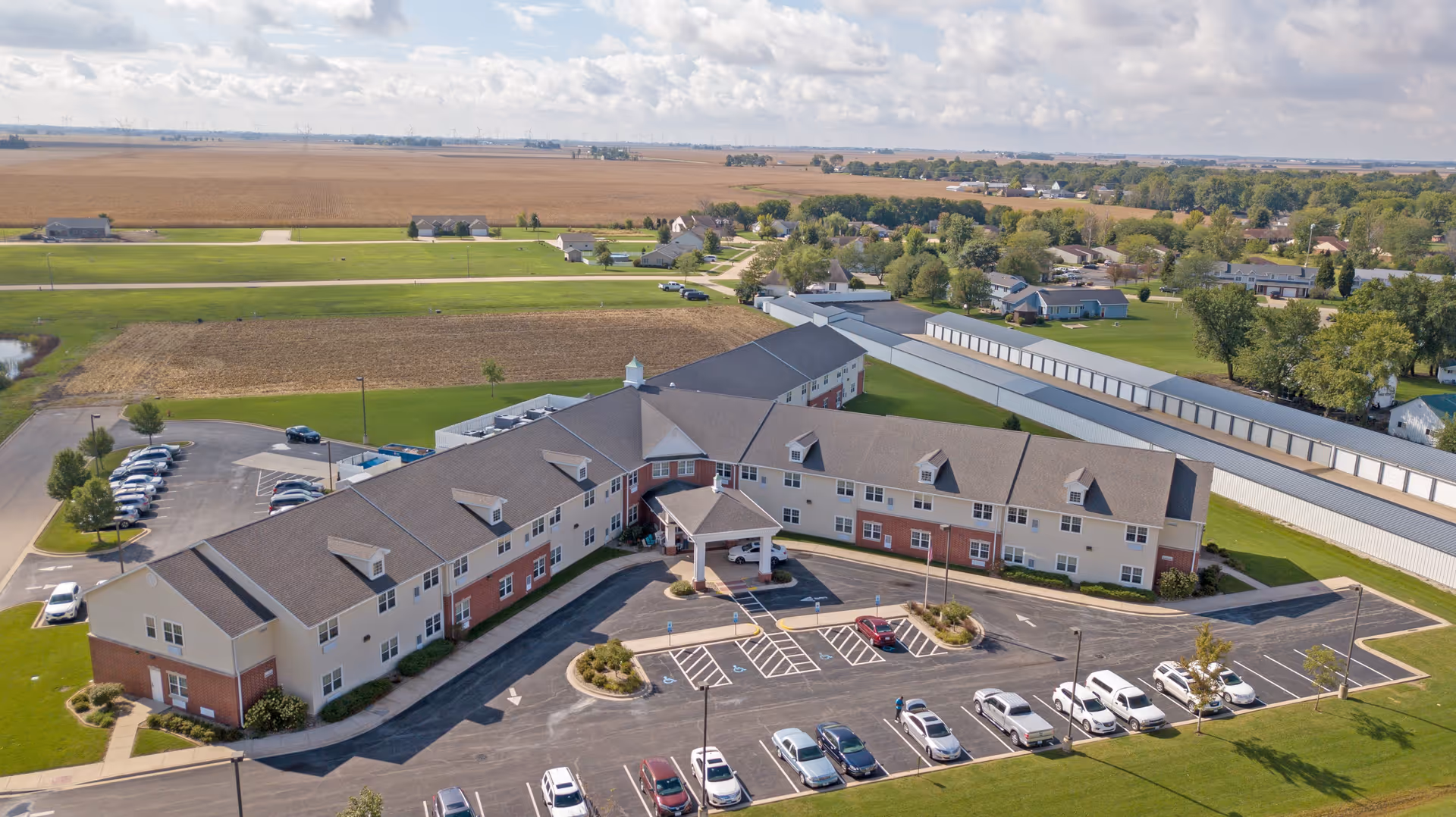 Aerial view of Heritage Woods of Dwight, a large senior living facility with a U-shaped building surrounded by parking lots and green lawns. The building has a beige and red brick exterior with multiple windows and a covered entrance. The surrounding area includes open fields, residential houses, and a long storage building.