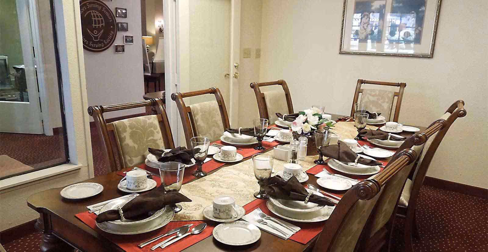 A dining room table set for eight people with elegant wooden chairs. The table is decorated with a beige table runner, red placemats, white china plates, cups, silverware, and brown cloth napkins with napkin rings. A floral centerpiece is placed in the middle of the table. The room has beige walls, a framed picture on the wall, and a window looking into another room.