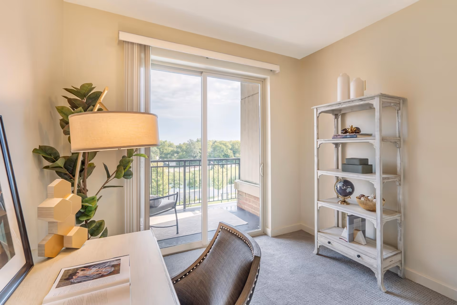 A bright room with a sliding glass door leading to a balcony with outdoor seating. Inside, there is a white wooden desk with a lamp, a plant, and an open book. To the right, there is a white distressed wooden shelving unit with decorative items. The room has beige walls and carpeted floor.