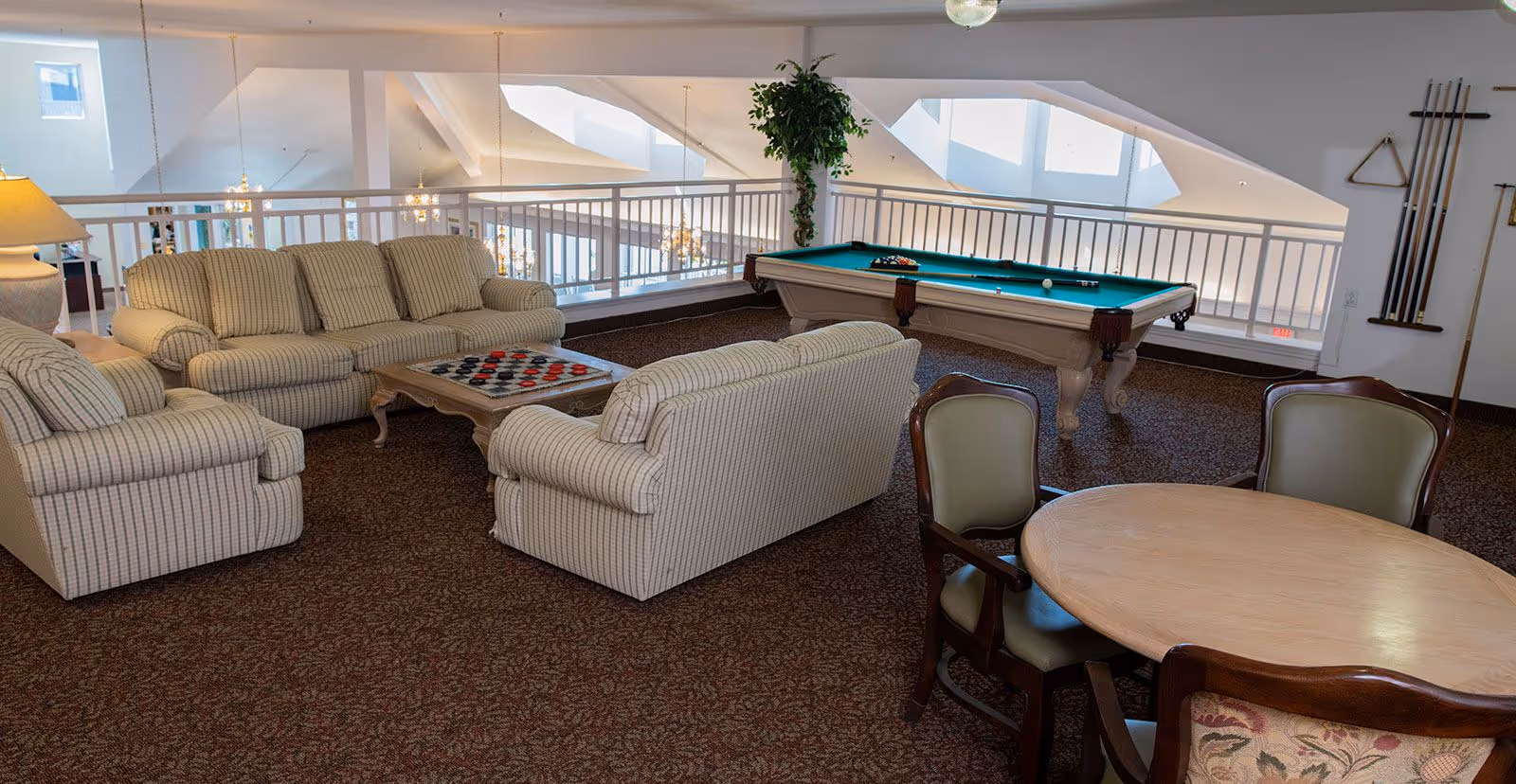A cozy recreational room with a beige striped sofa set, a wooden coffee table with a checkers game, a pool table with cues on the wall, a round wooden table with chairs, and a potted plant near a white railing overlooking a lower floor with chandeliers.