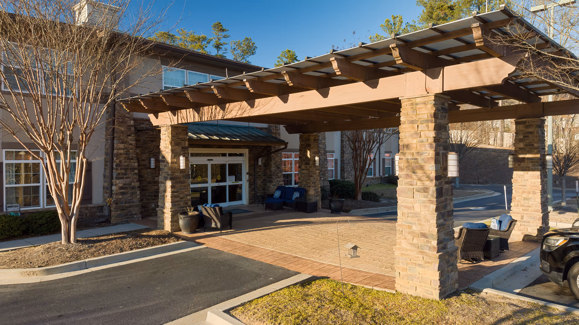 Entrance area of a senior living facility with a covered driveway supported by stone pillars, outdoor seating with chairs and small tables, leafless trees, and a building with large windows in the background under a clear sky.