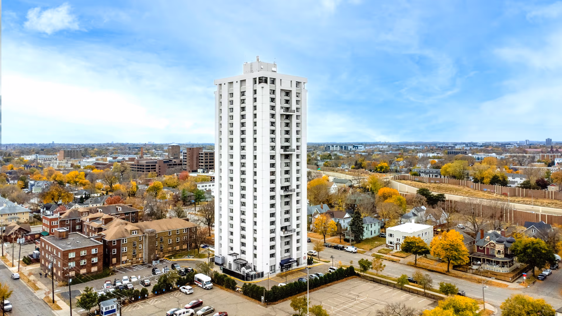 A tall white multi-story apartment building named Ebenezer Tower Apartments surrounded by a parking lot and residential neighborhood with trees showing autumn colors under a partly cloudy blue sky.