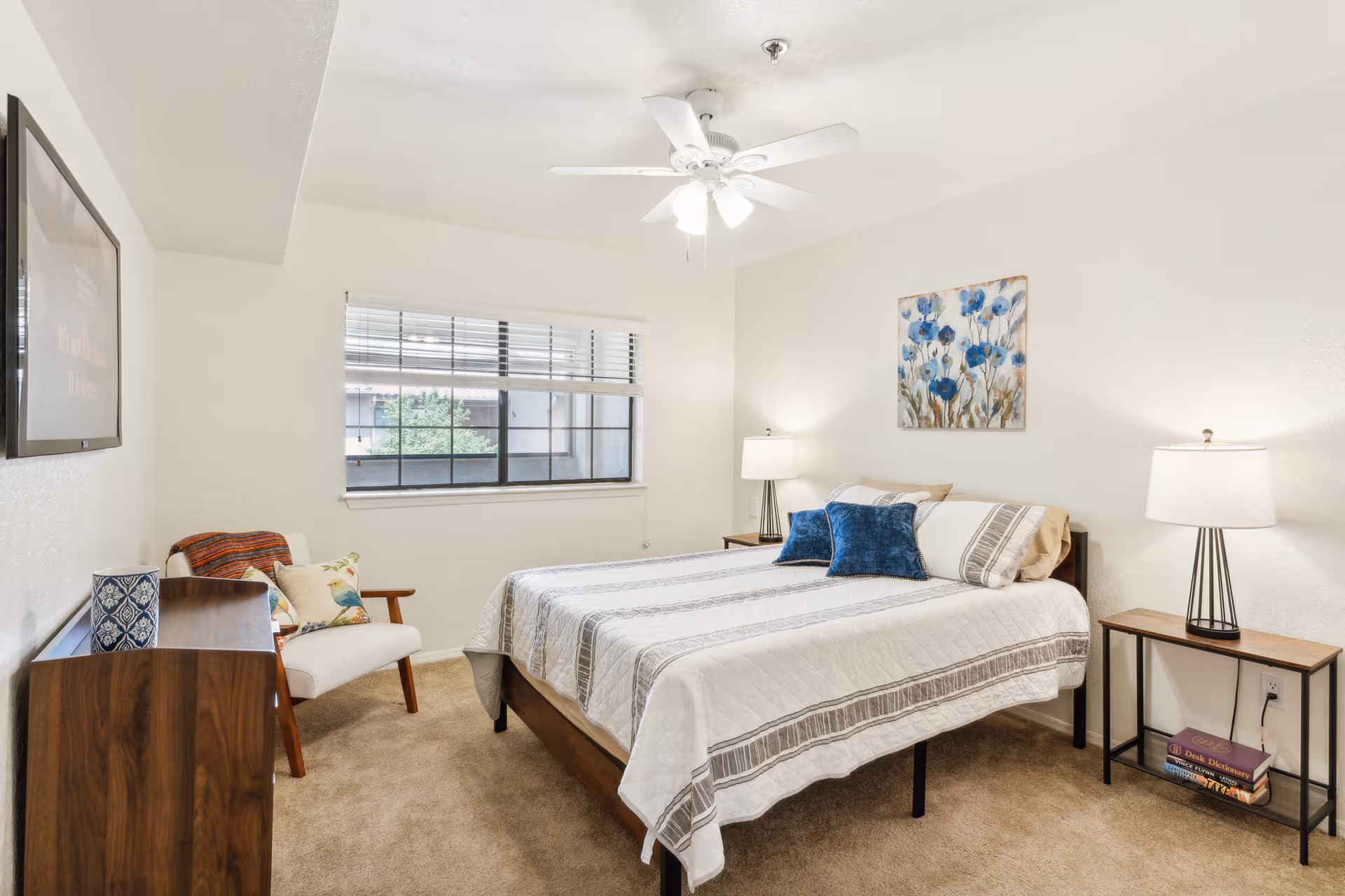 A bright bedroom with a large bed covered in a white and gray striped quilt, two blue accent pillows, and beige pillows. There are two bedside tables with matching lamps on either side of the bed. A colorful floral painting hangs above the bed. A window with blinds lets in natural light. A white armchair with decorative pillows and a wooden dresser with a blue patterned vase are also visible. The room has beige carpet and white walls with a ceiling fan overhead.