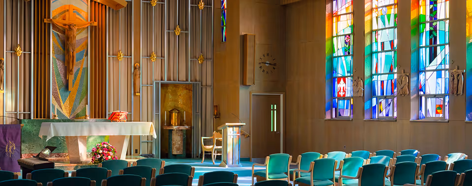 Interior view of a chapel with rows of teal chairs facing an altar. The altar is covered with a white cloth and decorated with flowers. Behind the altar is a large crucifix and colorful mosaic artwork. To the right, tall stained glass windows allow light to stream into the room. A lectern and a wooden chair are positioned near the altar.